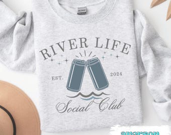 May include: Gray sweatshirt with the text "River Life Social Club" in a blue and white font. The design includes two blue cans clinking together, with waves below them. The text "Est. 2024" is also included.