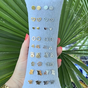 Könnte beinhalten: Ein grauer Samt-Präsentationskasten mit einer Auswahl an silbernen und goldenen Ohrsteckern. Die Ohrstecker haben verschiedene Designs, darunter Smileys, Herzen, Bienen, Blumen und Schmetterlinge.