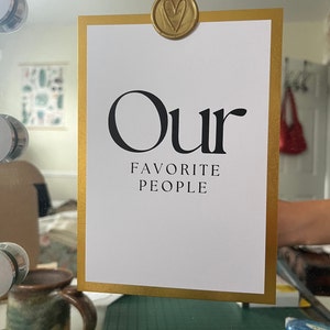 Op de afbeelding: Een witte kaart met een gouden lijst en de tekst "Our Favorite People" in zwarte letters. Een gouden, hartvormige zegel van was is bovenaan de kaart bevestigd.