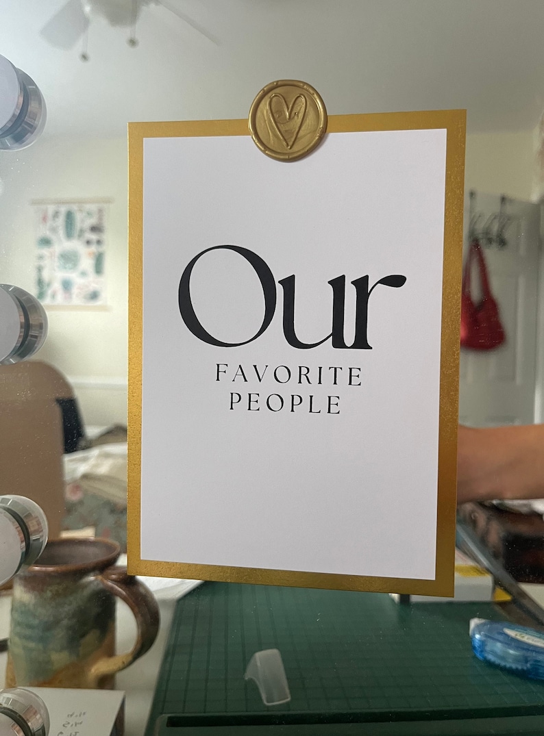 Op de afbeelding: Een witte kaart met een gouden lijst en de tekst "Our Favorite People" in zwarte letters. Een gouden, hartvormige zegel van was is bovenaan de kaart bevestigd.