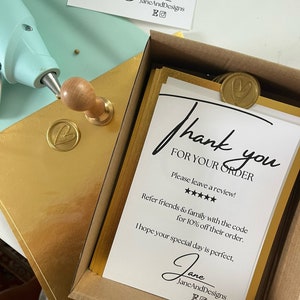 Op de afbeelding: Een witte kaart met zwarte tekst die zegt "Bedankt voor uw bestelling." De kaart zit in een bruine doos met gouden accenten. De kaart bevat ook een vijfsterrenbeoordeling en de tekst "Laat een beoordeling achter!" De kaart bevat ook de tekst "Verwijs vrienden en familie met de code voor 10% korting op hun bestelling." De kaart bevat ook de tekst "Ik hoop dat uw speciale dag perfect is." De kaart is ondertekend met "Jane JaneAndDesigns EO"