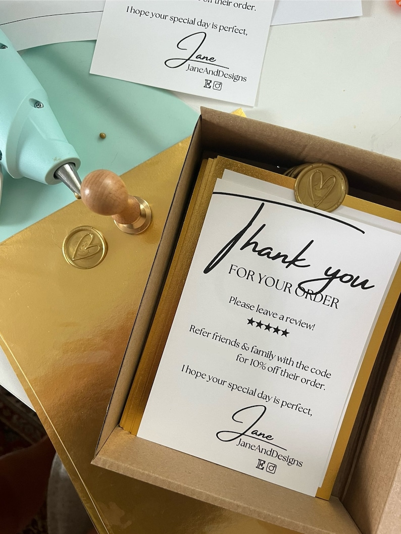Op de afbeelding: Een witte kaart met zwarte tekst die zegt "Bedankt voor uw bestelling." De kaart zit in een bruine doos met gouden accenten. De kaart bevat ook een vijfsterrenbeoordeling en de tekst "Laat een beoordeling achter!" De kaart bevat ook de tekst "Verwijs vrienden en familie met de code voor 10% korting op hun bestelling." De kaart bevat ook de tekst "Ik hoop dat uw speciale dag perfect is." De kaart is ondertekend met "Jane JaneAndDesigns EO"
