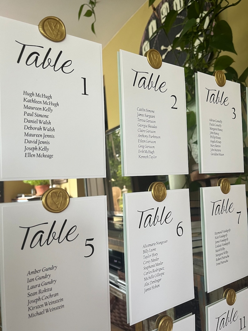 Op de afbeelding: Een set witte tafelkaarten met gouden waszegels en zwarte kalligrafie. De kaarten zijn genummerd van 1 tot en met 7 en 11. Elke kaart vermeldt namen voor een zitplaatskaart voor een bruiloftsreceptie.