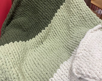 May include: A chunky knit blanket in shades of green and white. The blanket is divided into three sections: a dark olive green, a light sage green, and a white section. The texture is thick and plush, suggesting a cozy and warm feel.