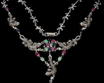 May include: A silver necklace with a pendant featuring a detailed lizard design. The pendant is adorned with green, red, and blue gemstones. The necklace is made of a delicate chain with lizard accents.