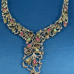 May include: A silver necklace with a delicate, intricate design featuring black, green, and pink gemstones. The necklace has a pendant that resembles a feather.
