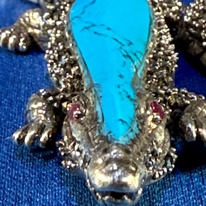 Può includere: Un ciondolo a forma di alligatore in argento con una pietra turchese sul dorso e occhi di rubino rosso. L'alligatore è dettagliato con una texture marmorea ed è posto su uno sfondo blu.