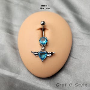 May include: A silver belly button ring featuring a blue gemstone and a heart-shaped charm with wings. The text "Model 1 blue / bleu" is visible. The text "Graf-O-Style" is in the lower right corner.