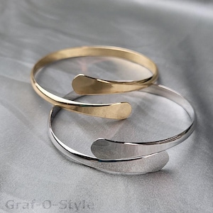 May include: Two simple bangle bracelets, one gold and one silver, with a slightly curved design. The bracelets are stacked on top of each other.