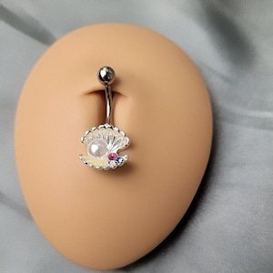 Peut inclure: Un piercing de nombril argenté avec un charme de perle et de coquille. La coquille est décorée de strass colorés.