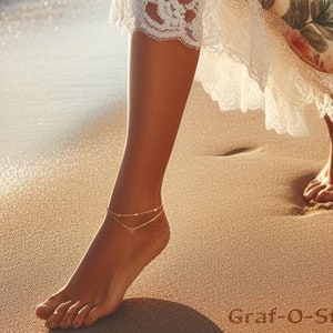 May include: A woman's ankle with two delicate gold anklets, one with a small heart charm. The anklets are on a bare foot on a sandy beach.