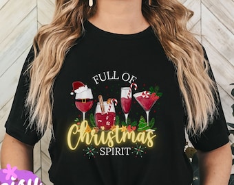 May include: Black t-shirt with a festive graphic. The design features wine glasses and cocktails with Christmas-themed decorations, and the text "FULL OF CHRISTMAS SPIRIT" in a glowing yellow font. The shirt is a crew neck.