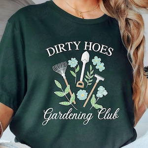 Può includere: Una maglietta verde scuro con il testo "Dirty Hoes Gardening Club" in bianco. Il design include una pala, un rastrello e una zappa con foglie verdi e fiori blu.