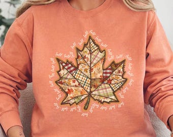 Pode incluir: Uma camisola coral com um grande desenho de folha. A folha é composta por vários patches xadrez e estampados em cores de outono. A palavra "fall" é repetida ao redor da borda da folha. A camisola tem mangas compridas e gola redonda.