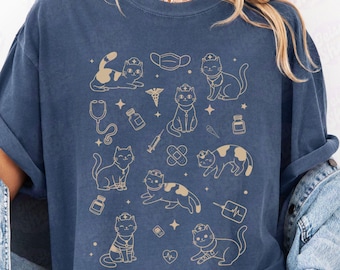 May include: Denim blue t-shirt featuring a repeating pattern of cartoon cats with medical symbols. The cats are wearing nurse hats and are surrounded by stethoscopes, syringes, and bandages. The word "Denim" is printed in a bold font at the bottom.