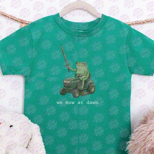 Op de afbeelding: Groen t-shirt met een cartoon kikker op een grasmaaier, met een zwaard. De tekst "we mow at dawn" staat onder de afbeelding. Het t-shirt hangt aan een houten hanger.