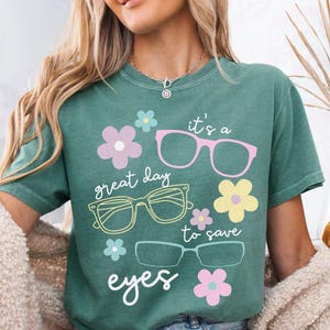 Może przedstawiać: Zielona koszulka z napisem "it's a great day to save eyes" oraz ilustracjami okularów i kwiatów w kolorze różowym, żółtym i niebieskim. Koszulka jest noszona z dżinsowymi szortami i kremowym kardiganem.