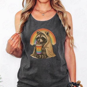 Könnte beinhalten: Dunkelgraues Tanktop mit einem Cartoon-Waschbären, der eine Kapuzen-Decke trägt und ein Getränk mit einem Strohhalm hält. Ein Regenbogen spannt sich hinter dem Waschbären. Der Waschbär ist das zentrale Element des Grafikdesigns.