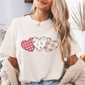 May include: Ivory-colored t-shirt featuring three heart appliqués. The hearts have different patterns: a red and pink gingham, a floral print, and a tan and white plaid. The word "Ivory" is written in a script font at the bottom.