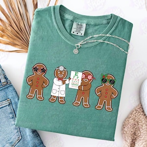 Può includere: Una maglietta Comfort Colors verde acqua con una grafica di quattro omini di pan di zenzero. Uno indossa occhiali da sole, un altro un camice da dottore e un altro tiene una tabella oculistica. Una collana d'argento con un ciondolo a forma di sole è drappeggiata sulla maglietta.