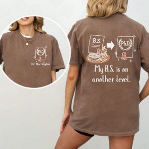 May include: Brown t-shirt with a white graphic on the front and back. The front graphic includes a PhD emblem and the name "Dr. Harrington." The back graphic shows a B.S. transforming into a PhD, with the text "My B.S. is on another level."