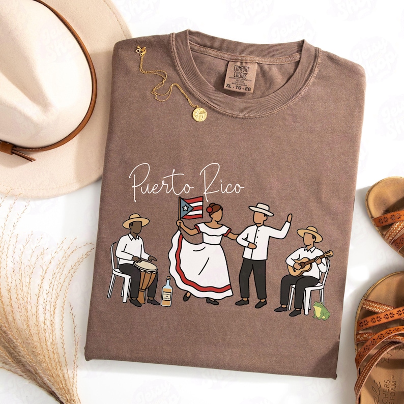 Pu&ograve; includere: Una maglietta marrone con la scritta "Puerto Rico" e una grafica di persone in abiti tradizionali. La grafica include una bandiera, un chitarrista e un batterista. Sono visibili anche una collana d'oro e un cappello.