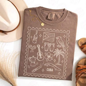 Puede incluir: Camiseta marrón con un gráfico blanco de iconos cubanos, incluyendo una bandera, una guitarra y una palmera. La camiseta tiene cuello redondo y una etiqueta que dice "XL - TG - EG". Un collar dorado y un sombrero están junto a la camiseta.