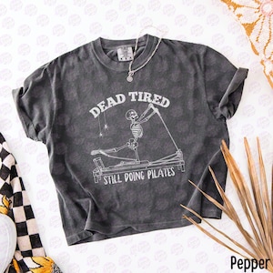 以下が含まれることがあります： チャコールグレーの半袖Tシャツ。リフォーマーマシンでピラティスをする骸骨の白いグラフィックが描かれています。骸骨の上には「DEAD TIRED」、下には「STILL DOING PILATES」という文字がかすれたフォントでプリントされています。
