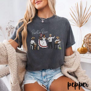 Pu&ograve; includere: T-shirt grigio scuro con la scritta "Puerto Rico" e un disegno di persone in abiti tradizionali, che suonano musica e tengono una bandiera. La maglietta &egrave; in stile casual.
