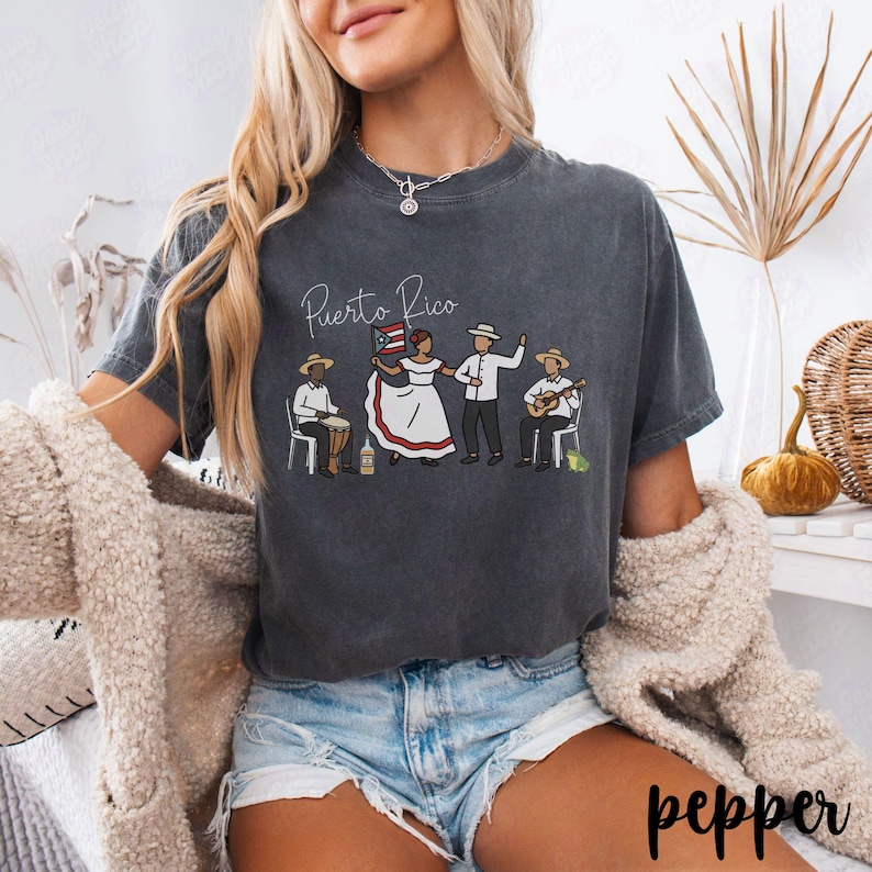Pu&ograve; includere: T-shirt grigio scuro con la scritta "Puerto Rico" e un disegno di persone in abiti tradizionali, che suonano musica e tengono una bandiera. La maglietta &egrave; in stile casual.