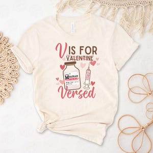 May include: A beige t-shirt with a graphic design that says "V is for Valentine Versed". The graphic features a bottle of medication labeled "Midazolam" and a syringe with hearts.