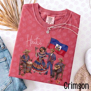 May include: Crimson-colored t-shirt featuring a graphic of Haitian people, a flag, and the word "Haiti." The design includes a drummer, a woman with maracas, a man holding the flag, and a guitarist. A silver necklace with a pendant is also visible.
