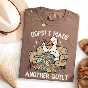 May include: Brown t-shirt featuring the text "OOPS! I MADE ANOTHER QUILT" and a cartoon duck sewing a quilt. The shirt has a crew neck and short sleeves. A gold necklace and a hat are also visible.
