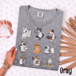 May include: A gray crew neck t-shirt featuring a graphic print of nine cartoon cats dressed as optometrists and ophthalmologists. The cats are shown interacting with various eye examination tools, including a phoropter, eye chart, and lens kit.