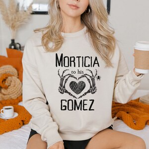 May include: A cream-coloured sweatshirt with the words "Morticia to his Gomez" in black. The design includes a heart made of skeleton hands and a spider. A coffee cup is held in the right hand.