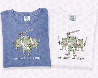 May include: Two t-shirts, one blue jean and one white, with a humorous frog wedding design. The bride frog is veiled and holds a sword, with two frog bridesmaids. Text reads "we bach at dawn".