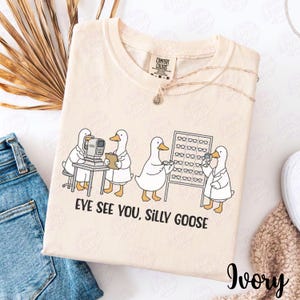 Może przedstawiać: Kremowa koszulka Comfort Colors z grafiką kreskówkowych kaczek w fartuchach laboratoryjnych, z napisem "EVE SEE YOU, SILLY GOOSE". Kaczki przedstawione są jako okuliści.