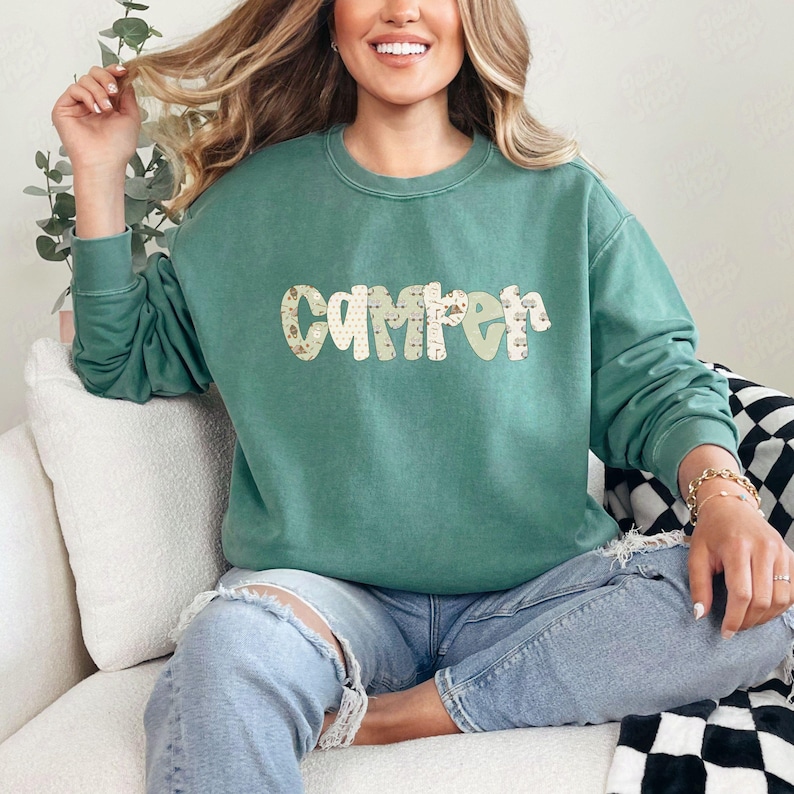 May include: A teal green sweatshirt with the word "CAMPER" in a decorative font. The letters have a floral pattern and a light green color. The sweatshirt is a crew neck style and is made of a soft material.