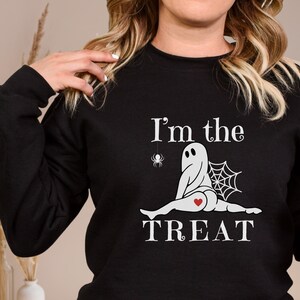 May include: A black long-sleeve sweatshirt featuring a white graphic of a ghost sitting with legs spread, a spider web, a small spider, and a red heart on the ghost's rear. The text "I'm the TREAT" is printed in white above and below the ghost graphic.