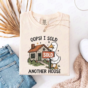 以下が含まれることがあります： クリーム色のTシャツ。家と「SOLD」の看板を持つガチョウのグラフィックが描かれています。Tシャツのテキストは「OOPS！I SOLD ANOTHER HOUSE」です。Tシャツは白い表面に折りたたまれています。