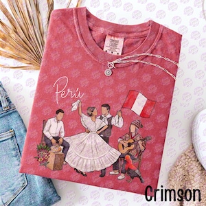 Op de afbeelding: Een rood, pigmentgeverfd T-shirt met ronde hals voorzien van een grafische illustratie van dansende en musicerende mensen, het woord "Perú" in wit schrift en een Peruaanse vlag. Een zilverkleurige ketting rust op de kraag.