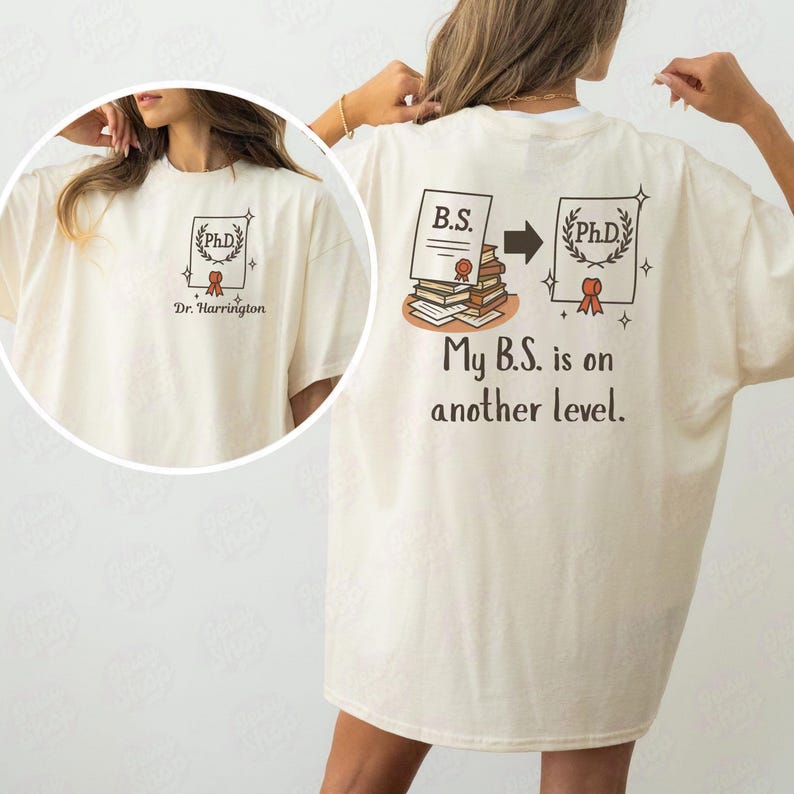 May include: Cream-colored t-shirt with a graphic on the front and back. The front graphic features a PhD certificate with the name "Dr. Harrington." The back graphic shows a B.S. certificate transforming into a PhD certificate with the text "My B.S. is on another level."