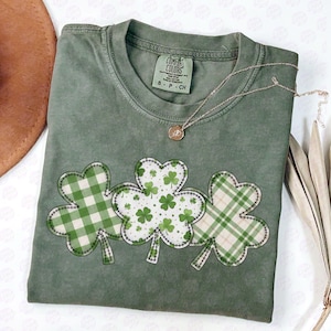 May include: A sage green t-shirt with three appliqued shamrocks in green and white plaid and floral patterns. A gold necklace with a pendant rests on the shirt. The shirt has a Comfort Colors tag.