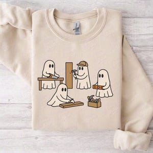Könnte beinhalten: Beiges Sweatshirt mit Rundhalsausschnitt und einem Cartoon-Motiv von vier Geistern, die mit Zimmermannswerkzeugen arbeiten. Ein Geist sägt Holz, ein anderer hämmert, ein dritter benutzt eine Wasserwaage und der letzte arbeitet mit einer Werkzeugkiste. Das Design ist in Schwarz und Braun gehalten.