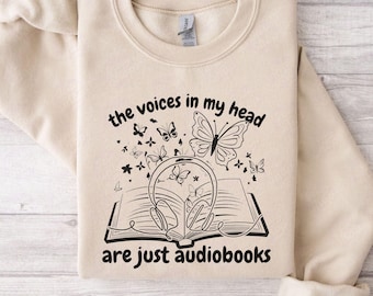 May include: A beige sweatshirt with the text "the voices in my head are just audiobooks" printed on the front. The text is surrounded by a design of a book with headphones and butterflies.