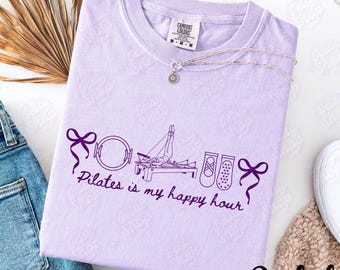 Puede incluir: Una camiseta de color lila claro con la frase "Pilates is my happy hour" en escritura morada oscura. La camiseta presenta un gráfico de equipo de Pilates, zapatillas de ballet y lazos. Un collar plateado con un colgante de sol está sobre la camiseta.