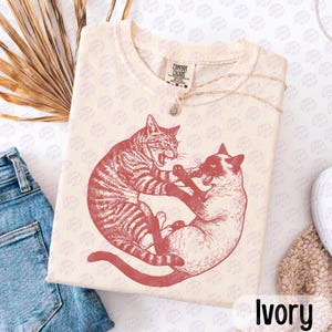 Könnte beinhalten: Ein cremefarbenes Kurzarm-T-Shirt mit einem dunkelroten Grafikdruck von zwei Katzen, die in kreisförmiger Anordnung spielen oder kämpfen. Das T-Shirt hat einen Rundhalsausschnitt und ein Comfort Colors Markenetikett am inneren Kragenbereich.
