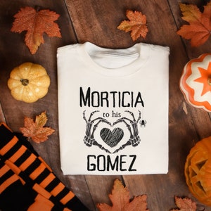 May include: A cream-coloured t-shirt with the words "MORTICIA to his GOMEZ" in black, featuring a heart formed by skeleton hands. The shirt is surrounded by autumn decorations, including pumpkins and leaves.