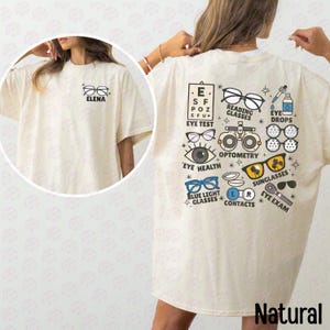May include: A cream-coloured t-shirt featuring a small graphic on the front chest with glasses and the name ELENA. The back displays various optometry-themed illustrations including an eye chart, reading glasses, eye drops, and eye exam tools.