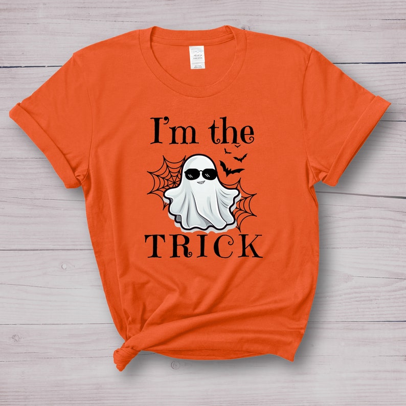May include: An orange short-sleeved t-shirt featuring a graphic of a white ghost wearing black sunglasses. The design includes black spiderwebs, small bats, and the text "I'm the TRICK" printed in a black serif font centered on the chest.